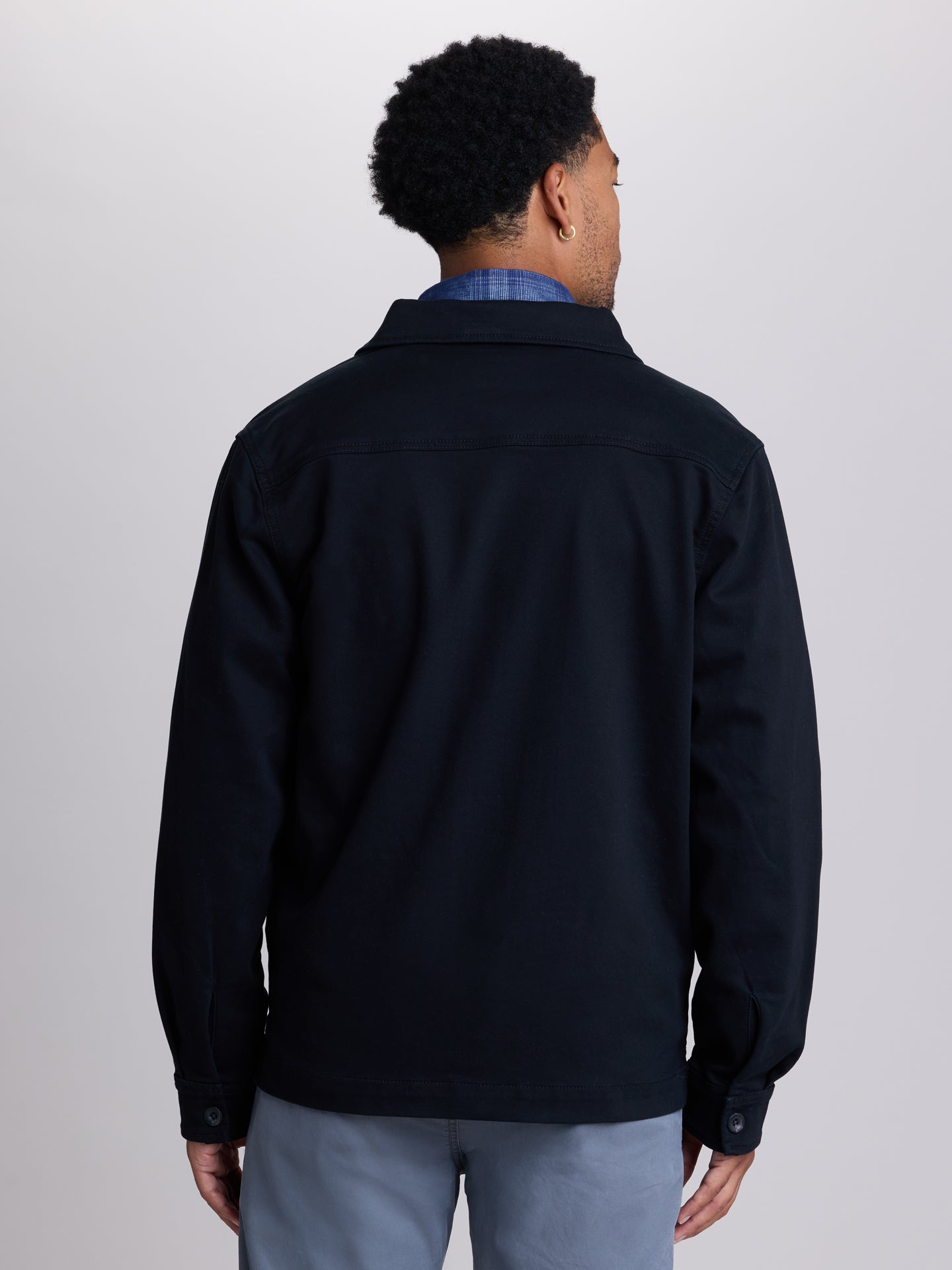 A man viewed from the back wearing a dark navy blue jacket over a blue plaid shirt and light gray trousers, set against a plain light gray studio background.