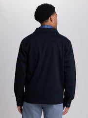 A man viewed from the back wearing a dark navy blue jacket over a blue plaid shirt and light gray trousers, set against a plain light gray studio background.