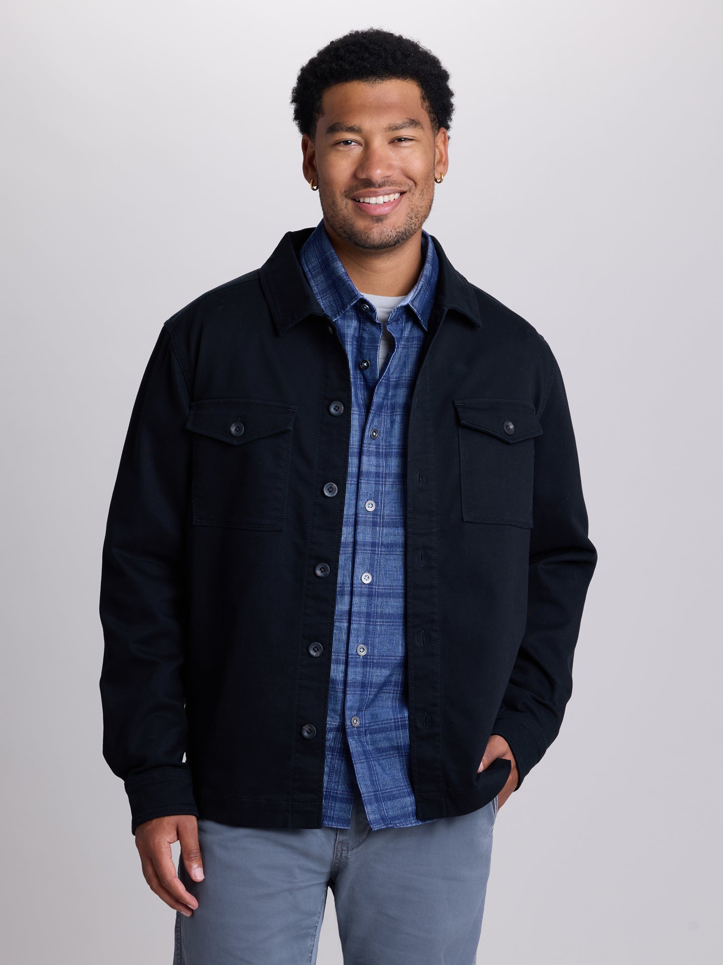 A man wearing a black utility jacket over a blue plaid button-down shirt and light gray jeans, standing against a light gray studio background | Black
