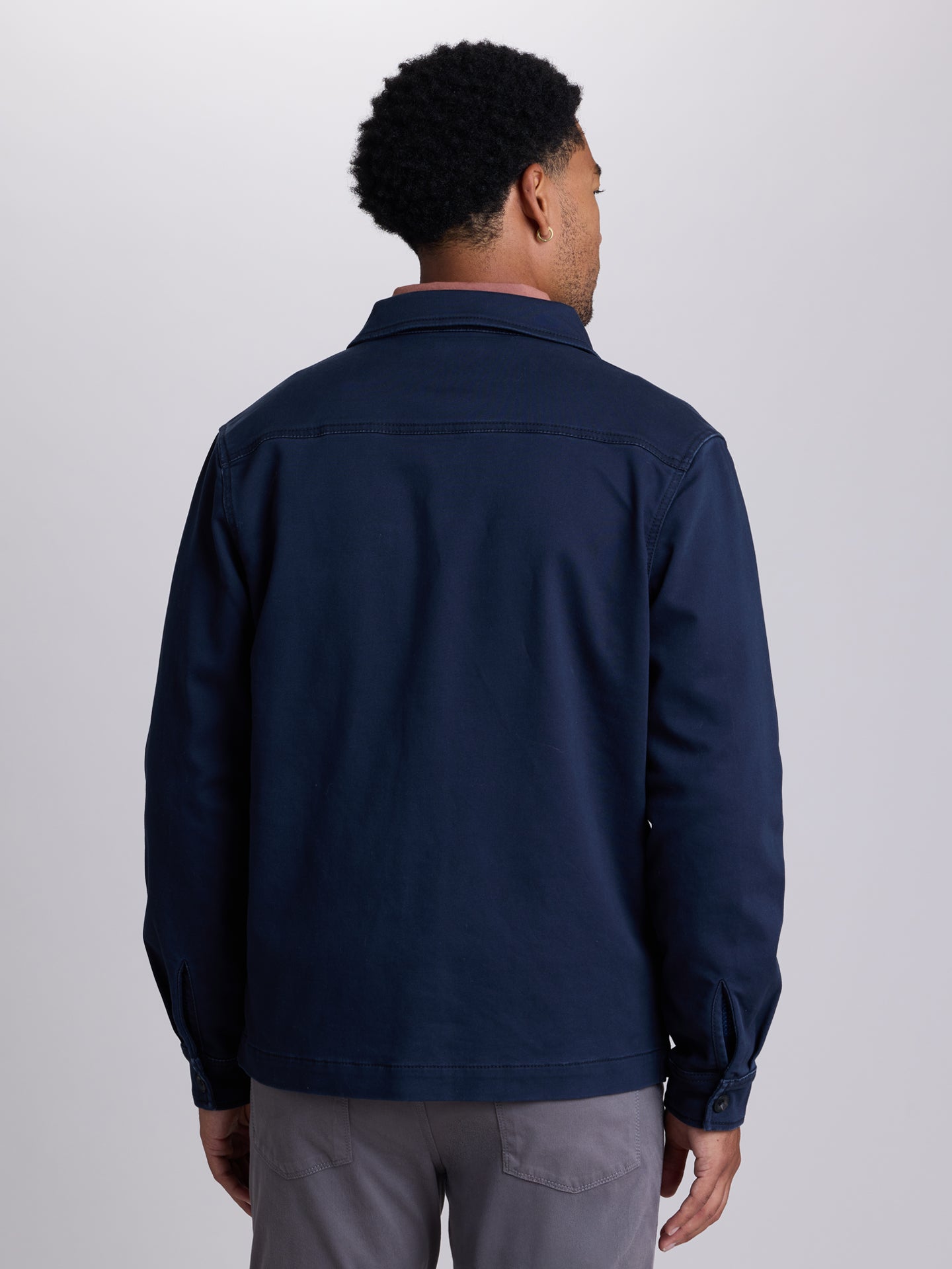 A man viewed from the back wearing a dark navy blue long-sleeve utility jacket and gray trousers, standing against a light gray studio background. | Twilight