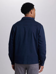 A man viewed from the back wearing a dark navy blue long-sleeve utility jacket and gray trousers, standing against a light gray studio background. | Twilight