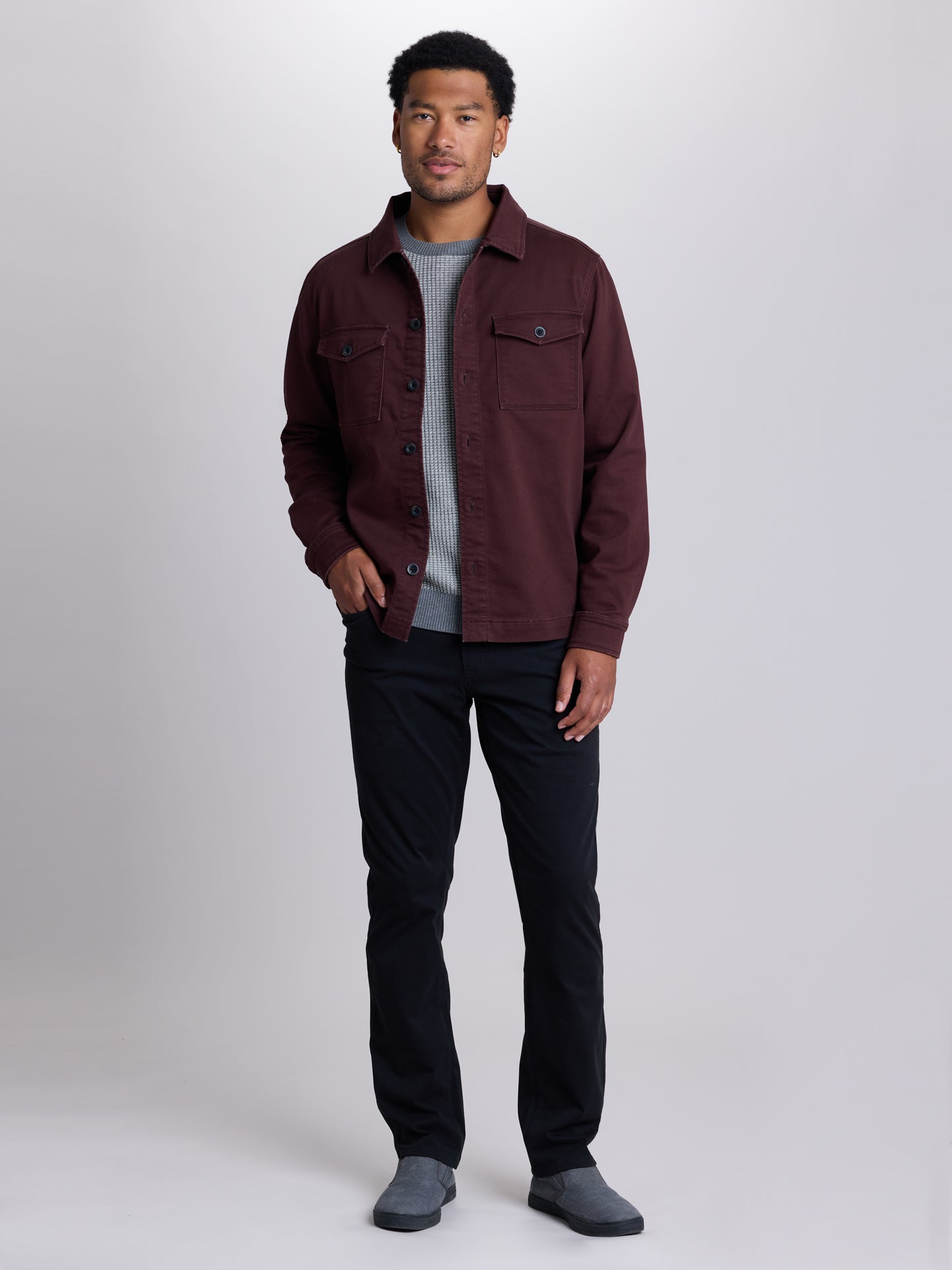 A man modeling a dark burgundy button-up overshirt over a gray knit sweater, black pants, and gray slip-on shoes, photographed against a light gray studio background. | Dark Oxblood