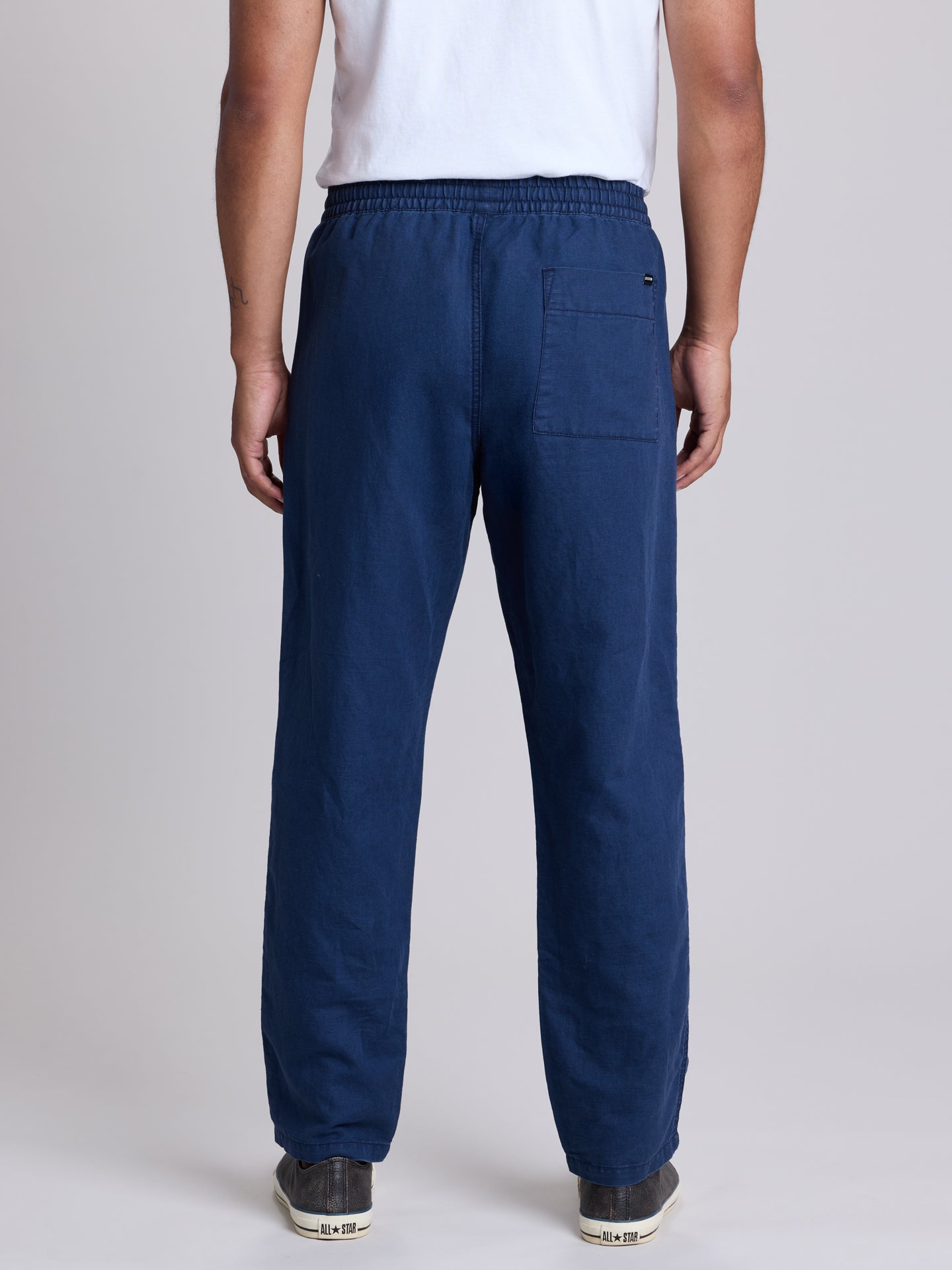 A man viewed from the back wearing navy blue relaxed-fit pants, a white t-shirt, and dark sneakers against a light gray studio background. | Deep Blue