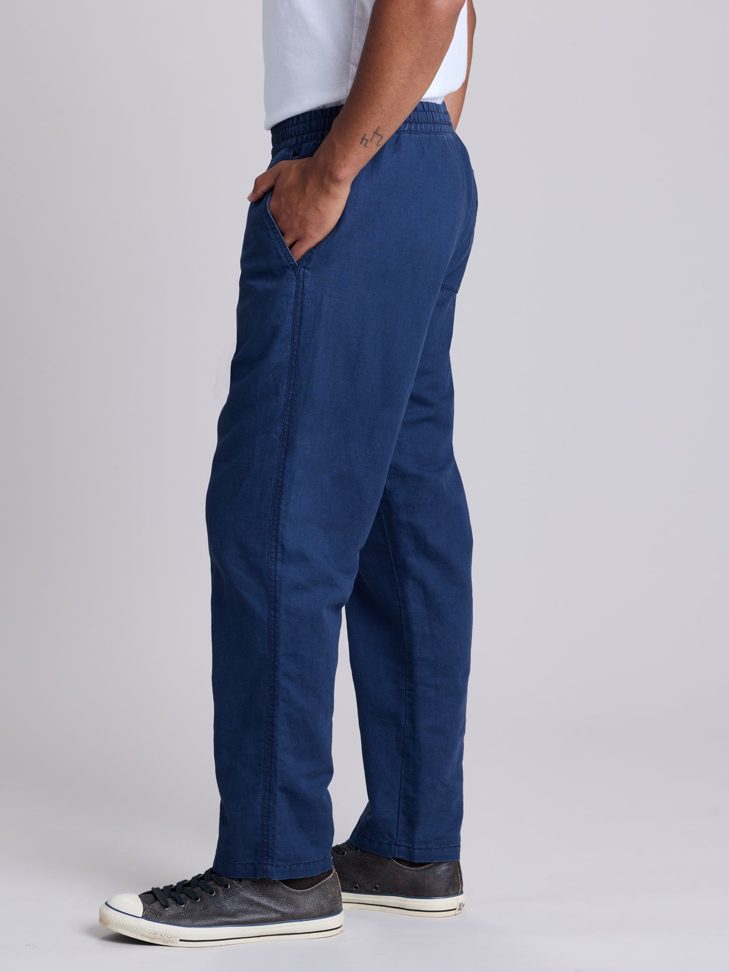 A person shown from the waist down wearing navy blue straight-leg pants, a white t-shirt, and dark gray sneakers against a light gray studio background. | Deep Blue