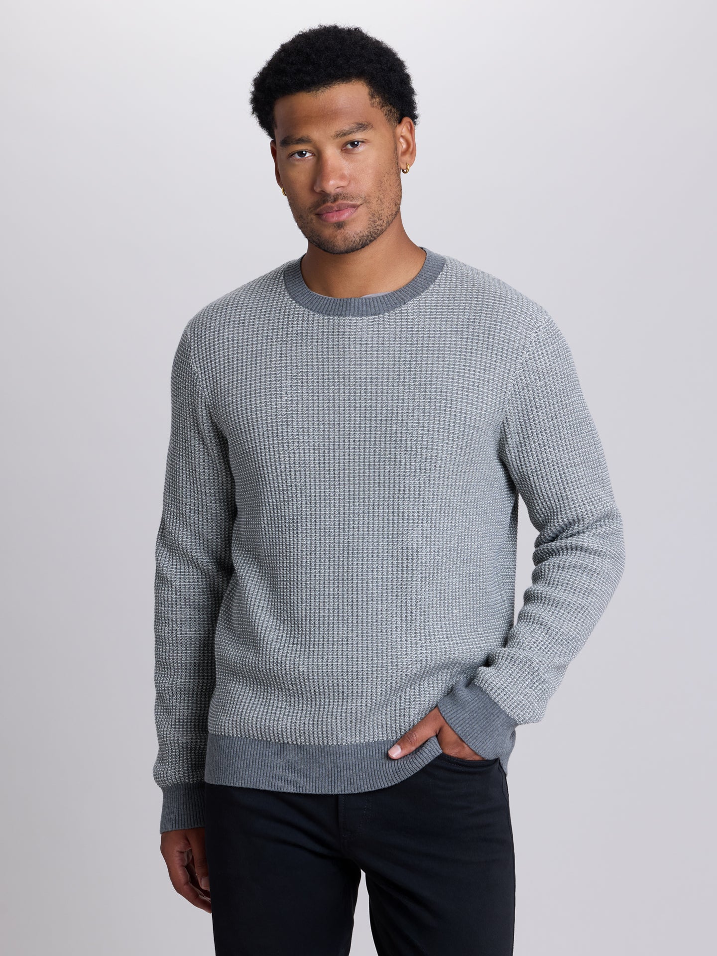 A man wearing a gray waffle-knit crewneck sweater and dark pants, standing against a light gray studio background. | Dark Medium Grey Heather