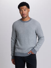 A man wearing a gray waffle-knit crewneck sweater and dark pants, standing against a light gray studio background. | Dark Medium Grey Heather