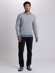 A man modeling a light gray ribbed knit crewneck sweater with darker gray ribbed trim at the collar, cuffs, and hem, paired with dark trousers and gray slip-on shoes, against a light gray studio background.  | Dark Medium Grey Heather