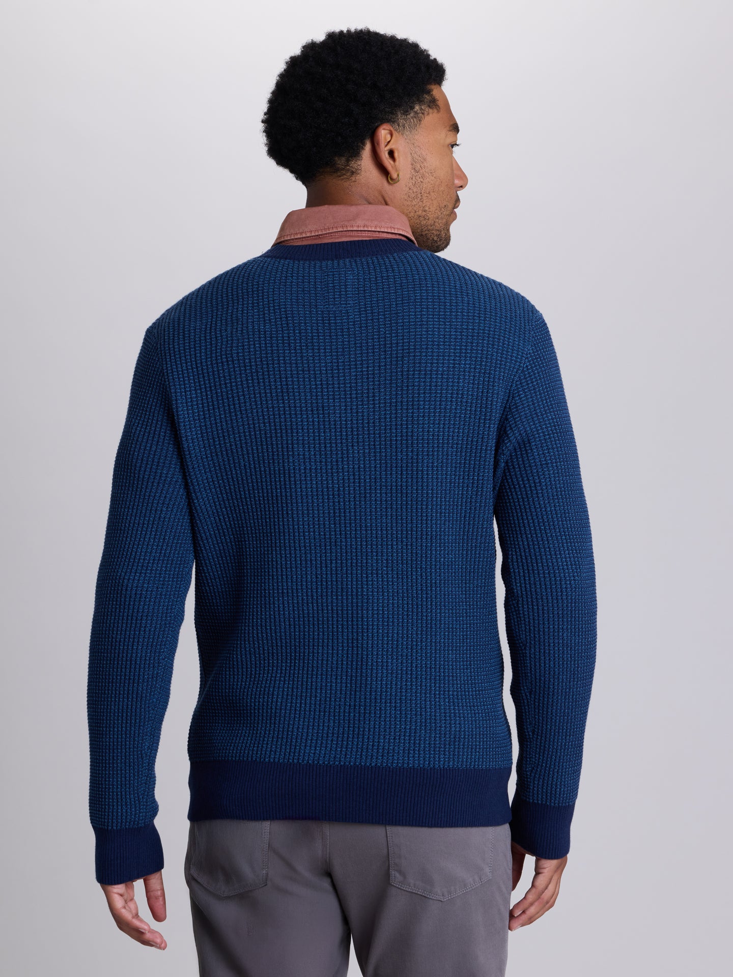 A man viewed from the back wearing a textured navy blue knit sweater over a dusty rose collared shirt, paired with gray trousers, against a light gray studio background.  | Navy