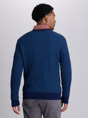 A man viewed from the back wearing a textured navy blue knit sweater over a dusty rose collared shirt, paired with gray trousers, against a light gray studio background.  | Navy
