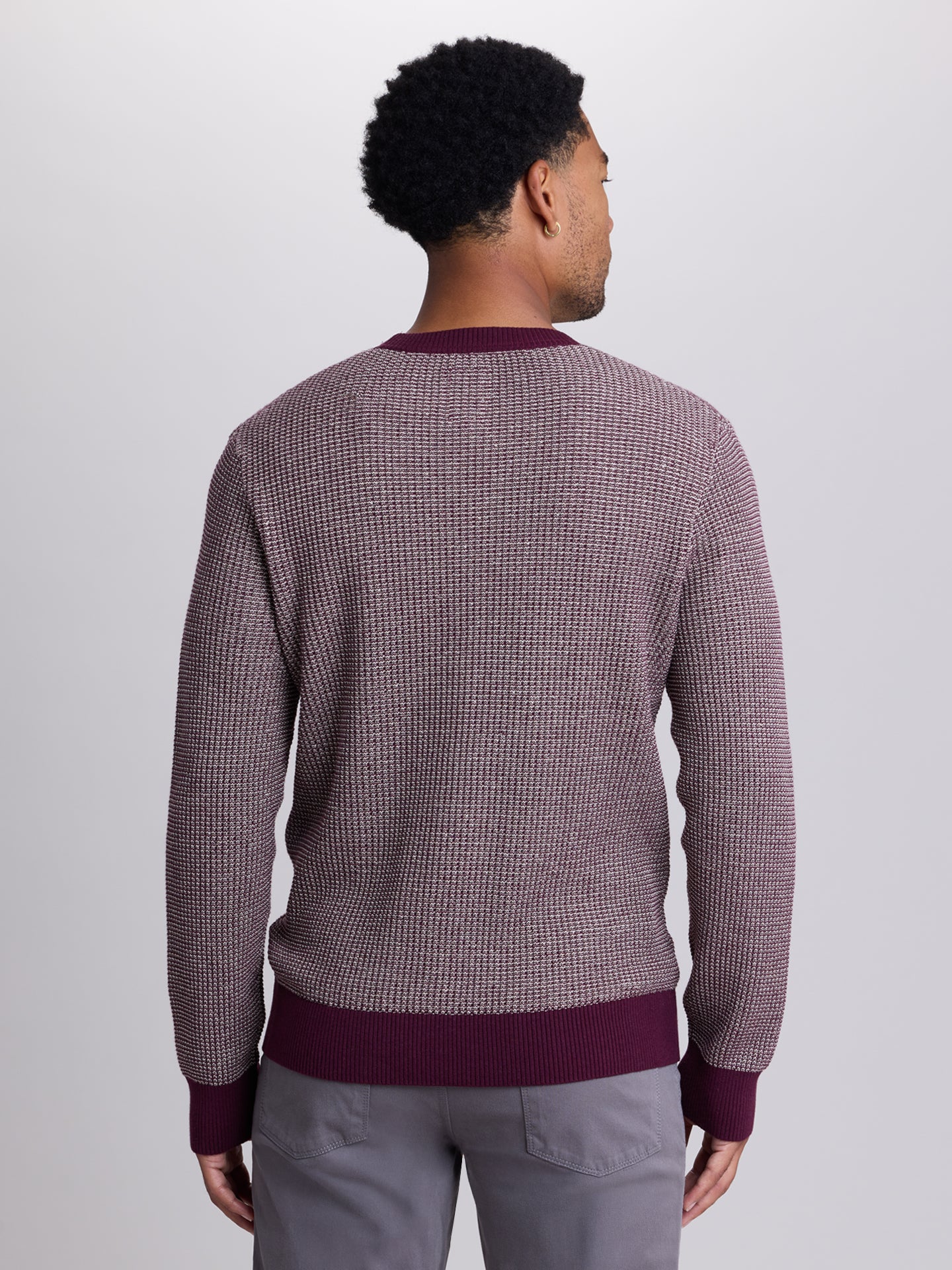 A man viewed from behind wearing a textured waffle-knit sweater with a small-scale check pattern in dusty purple and white, featuring solid dark purple ribbed trim at the collar, cuffs, and hem, paired with gray trousers. | Cloak 