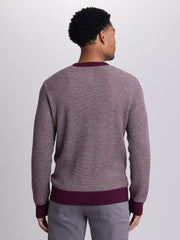 A man viewed from behind wearing a textured waffle-knit sweater with a small-scale check pattern in dusty purple and white, featuring solid dark purple ribbed trim at the collar, cuffs, and hem, paired with gray trousers. | Cloak 