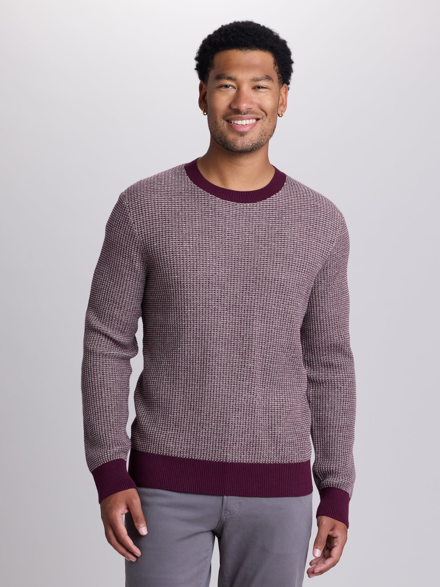 A man wearing a textured waffle-knit crewneck sweater in a dusty mauve-purple color with contrasting dark burgundy ribbed trim at the collar, cuffs, and hem, paired with gray pants against a light gray studio background. | Cloak 