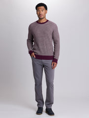 A man modeling a ribbed knit crewneck sweater in a dusty mauve-purple color with contrasting dark burgundy trim at the collar, cuffs, and hem, paired with gray trousers and dark blue shoes, against a light gray studio background. | Cloak 