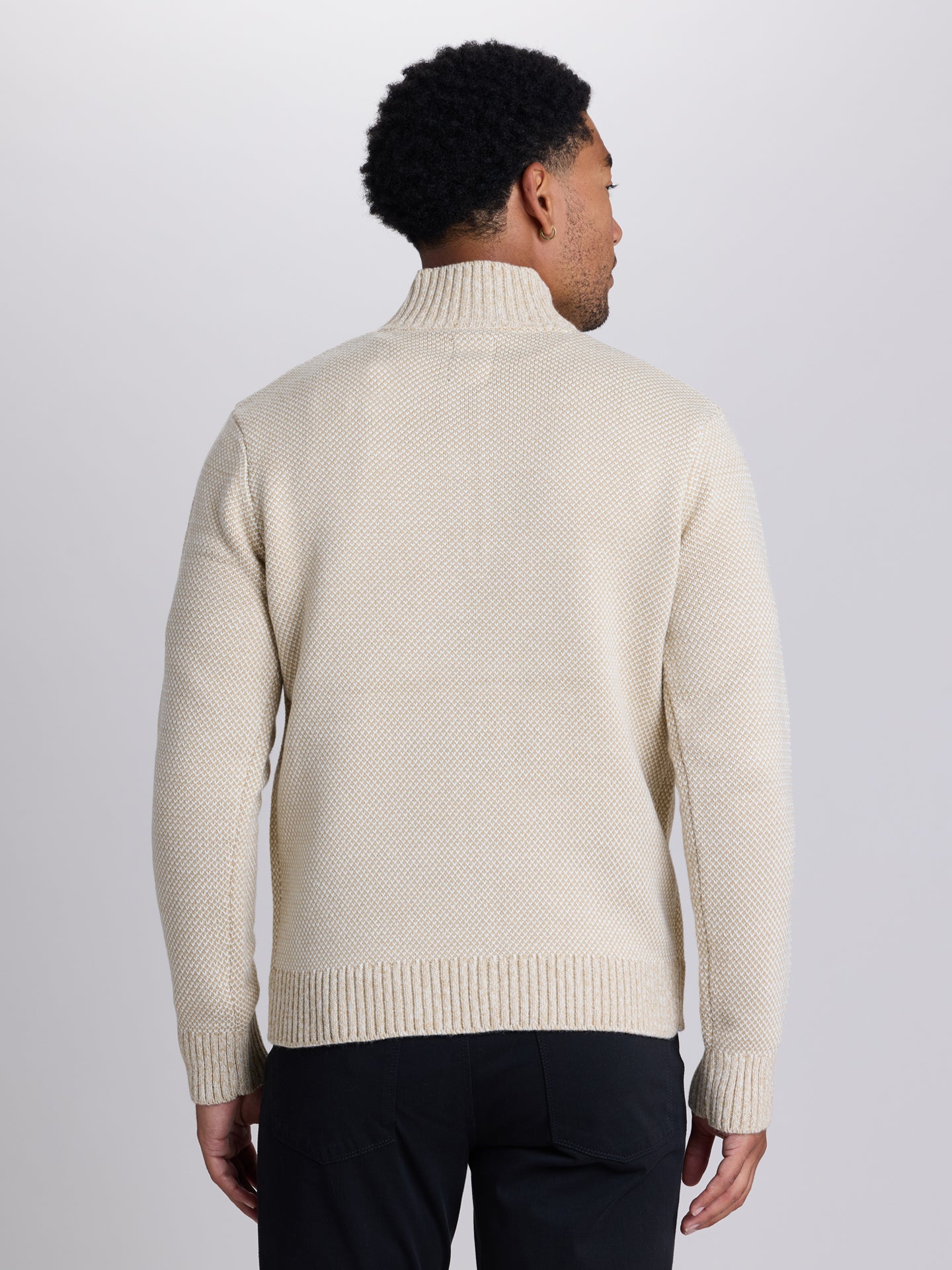 A man viewed from the back wearing a cream-colored knit mock neck sweater and black trousers, set against a plain light gray studio background. | Chai
