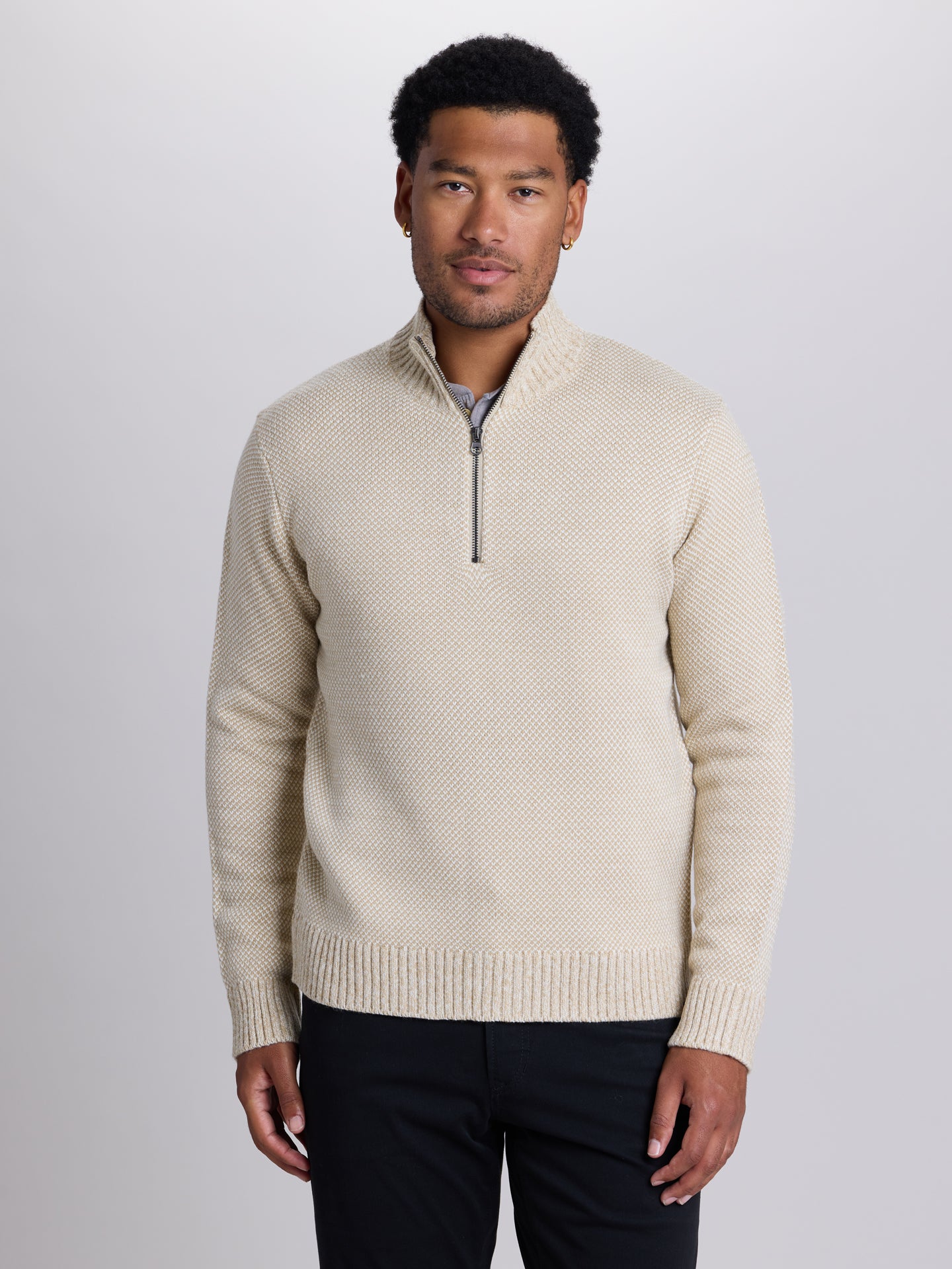 A man wearing a cream-colored textured knit quarter-zip sweater with a stand collar, paired with dark trousers, photographed against a neutral light gray studio background. | Chai