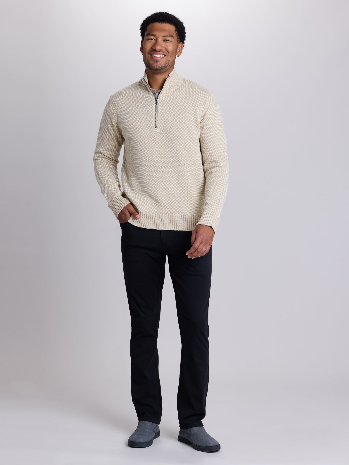 A man wearing a cream-colored quarter-zip knit sweater, black trousers, and gray slip-on shoes, standing against a light gray studio background. | Chai