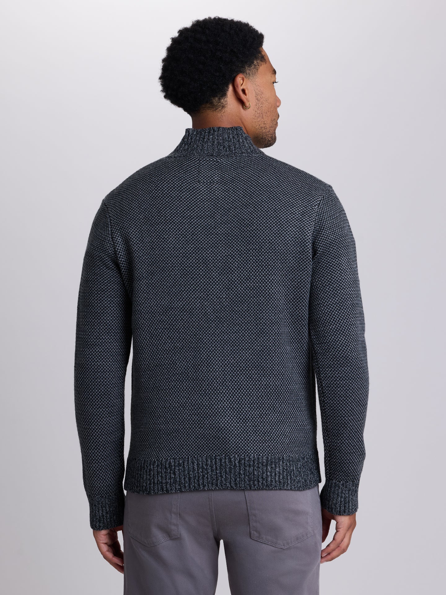 A man viewed from the back wearing a dark charcoal gray textured knit mock neck sweater and gray trousers, set against a plain light gray studio background. | Skillet