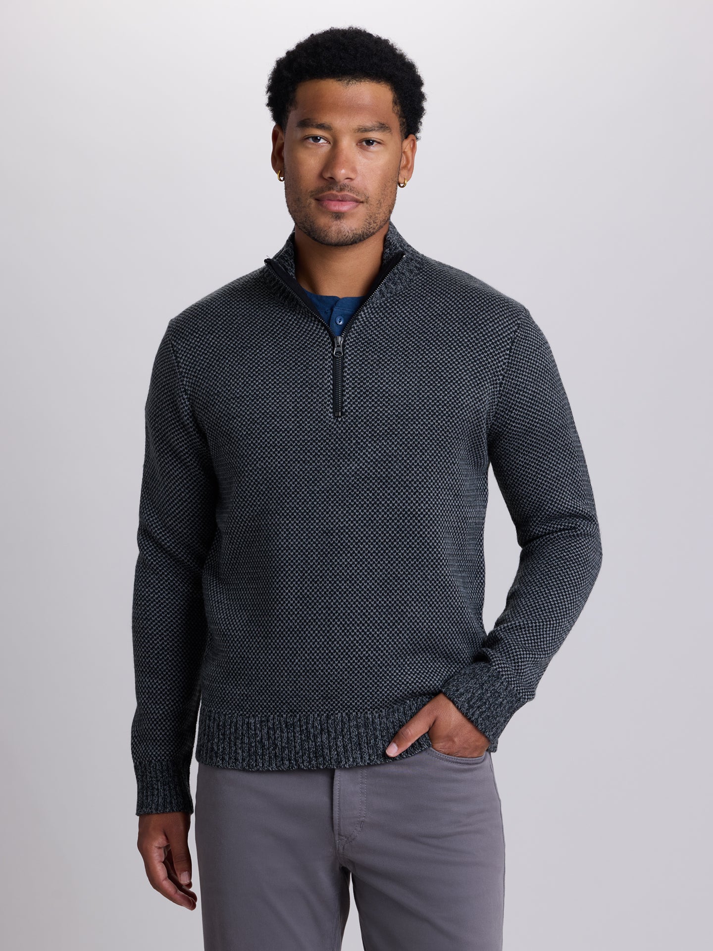 A man wearing a dark gray textured knit quarter-zip sweater over a blue undershirt and gray pants, standing against a light gray studio background. | Skillet