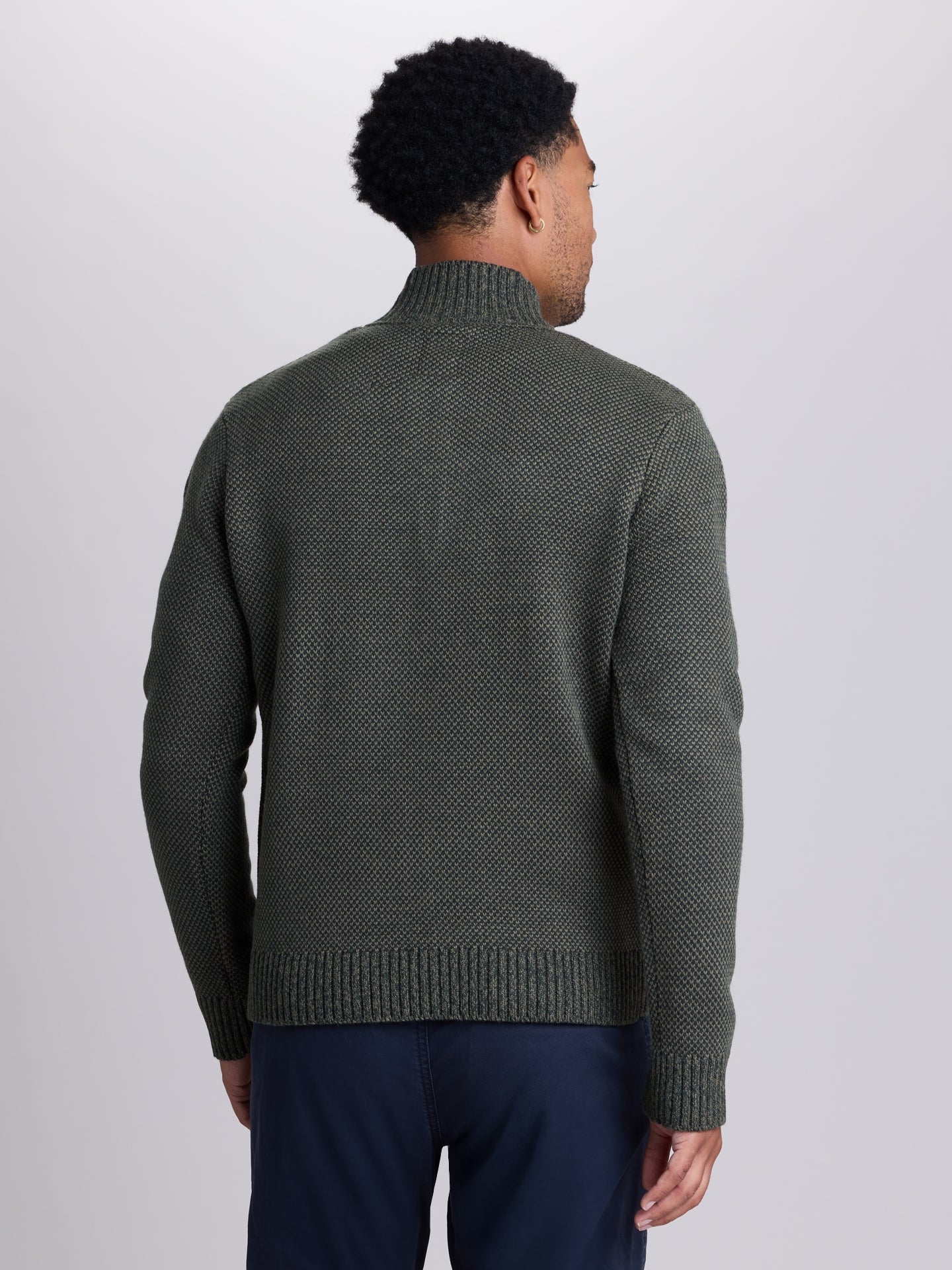 A man viewed from the back wearing a textured dark olive green mock-neck sweater and navy blue trousers, set against a plain light gray studio background.  | Dark Spruce