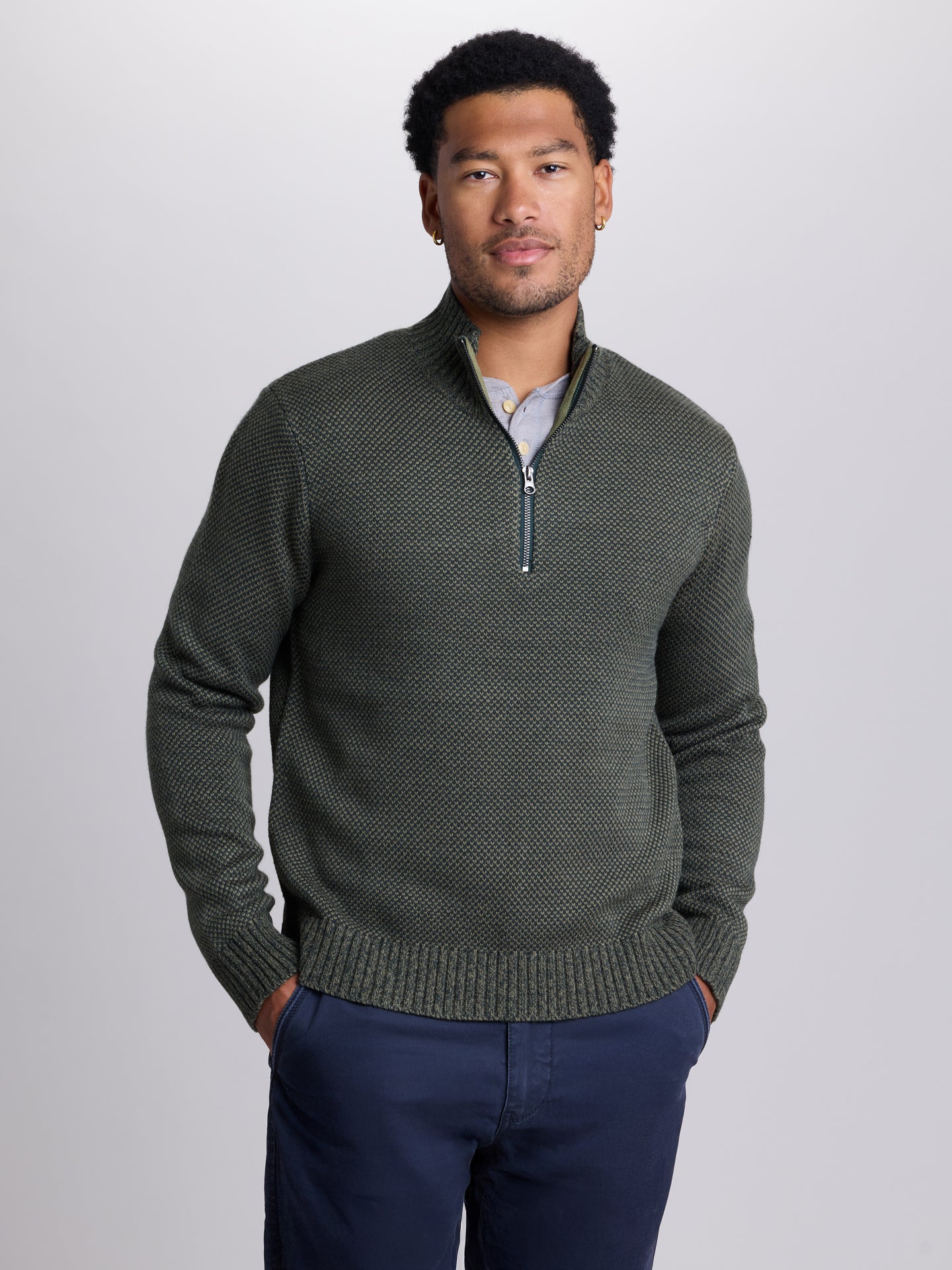 A man wearing a dark olive green textured knit quarter-zip sweater over a light blue collared shirt, paired with navy blue trousers, standing against a light gray studio background. | Dark Spruce