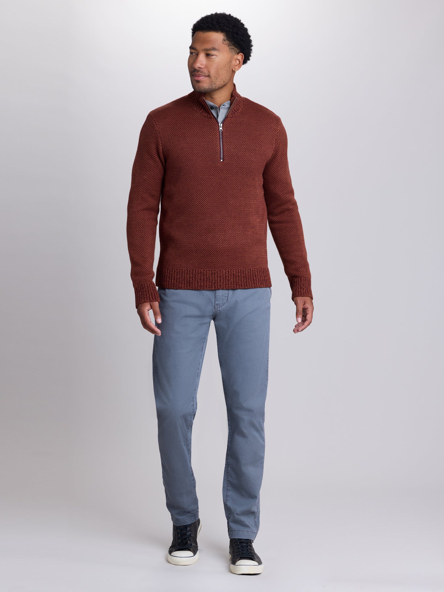 A man modeling a rust-brown half-zip knit sweater, paired with light blue trousers, a light blue collared shirt, and black sneakers, against a neutral gray studio background. | Redwood