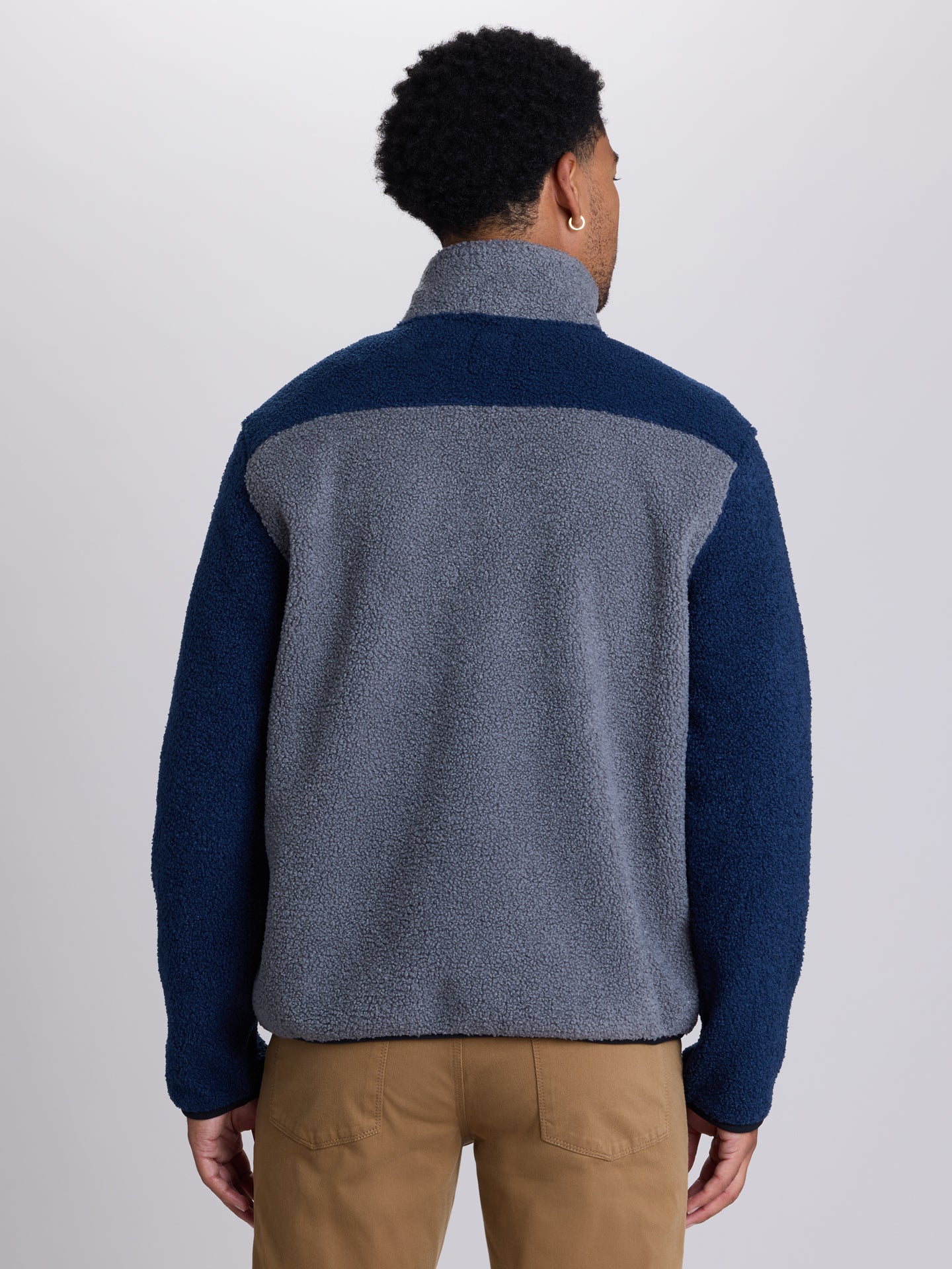 A man viewed from the back wearing a color-block fleece pullover with a gray body and navy sleeves, paired with tan pants, against a light gray studio background. | Charcoal