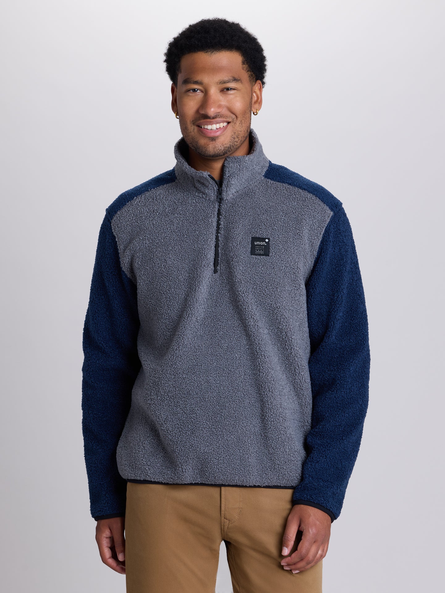 A man wearing a color-blocked quarter-zip fleece pullover with a heather gray body and navy sleeves, paired with tan pants, against a light gray studio background. | Charcoal