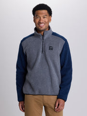 A man wearing a color-blocked quarter-zip fleece pullover with a heather gray body and navy sleeves, paired with tan pants, against a light gray studio background. | Charcoal
