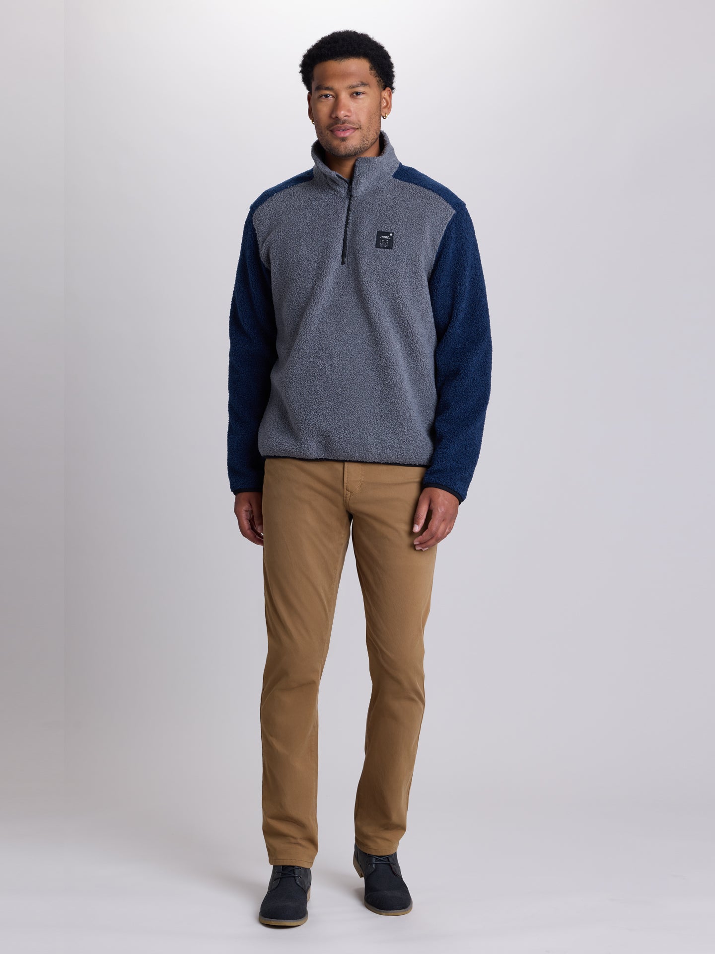 A man modeling a quarter-zip fleece pullover sweater with heather gray body and navy blue sleeves, paired with tan pants and dark shoes, against a light gray studio background. | Charcoal