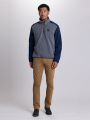 A man modeling a quarter-zip fleece pullover sweater with heather gray body and navy blue sleeves, paired with tan pants and dark shoes, against a light gray studio background. | Charcoal