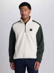 A man wearing a fleece pullover sweater with a cream-colored body and dark green sleeves, featuring a quarter-zip collar and a small black logo patch on the left chest, set against a plain light gray studio background. | Powder