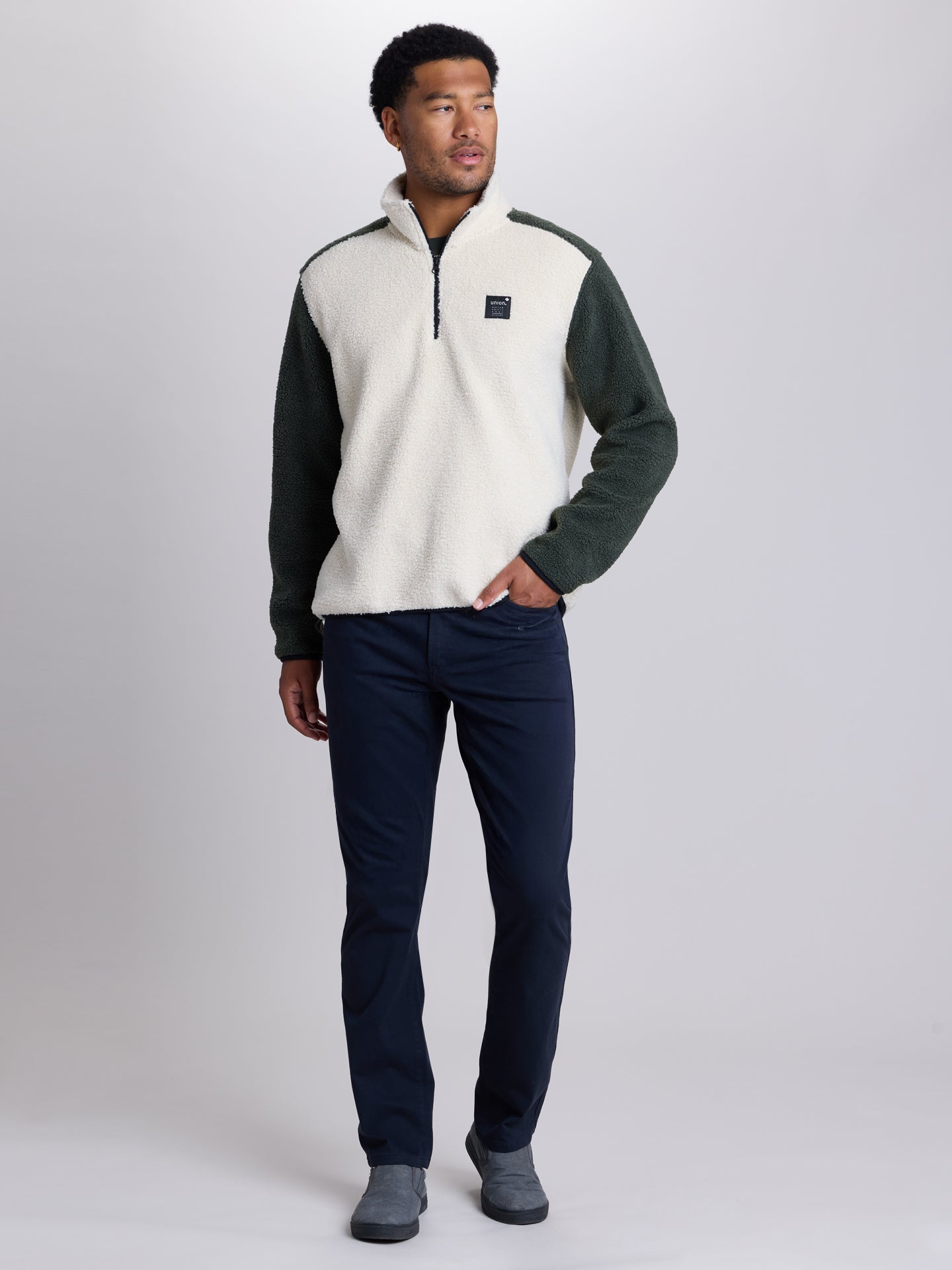 A man modeling a quarter-zip fleece pullover sweater with a cream body and dark green sleeves, paired with black pants and gray shoes, against a light gray studio background. | Black