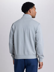 A man viewed from the back wearing a light gray quarter-zip sweatshirt and dark blue pants, standing against a plain light gray studio background. | Cobra