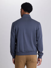 A man viewed from the back wearing a slate blue quarter-zip sweatshirt and tan pants, set against a plain light gray studio background. | Forged