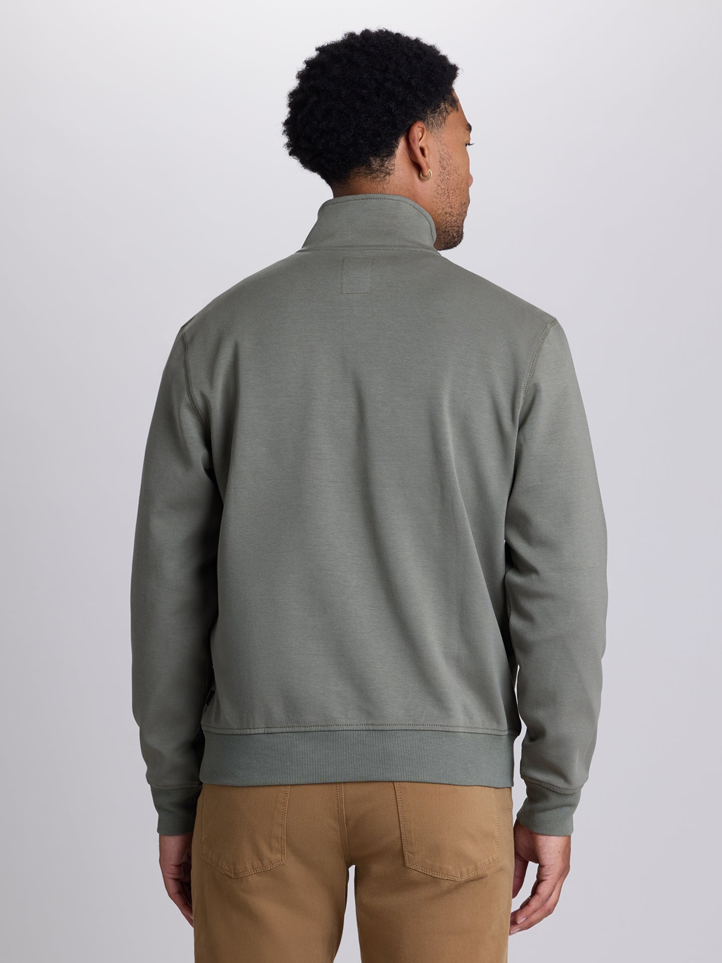 A man viewed from the back wearing a muted sage green quarter-zip pullover sweatshirt and tan pants, set against a plain light gray studio background. | Dusty Olive