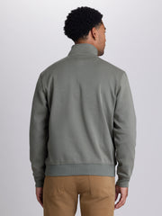 A man viewed from the back wearing a muted sage green quarter-zip pullover sweatshirt and tan pants, set against a plain light gray studio background. | Dusty Olive