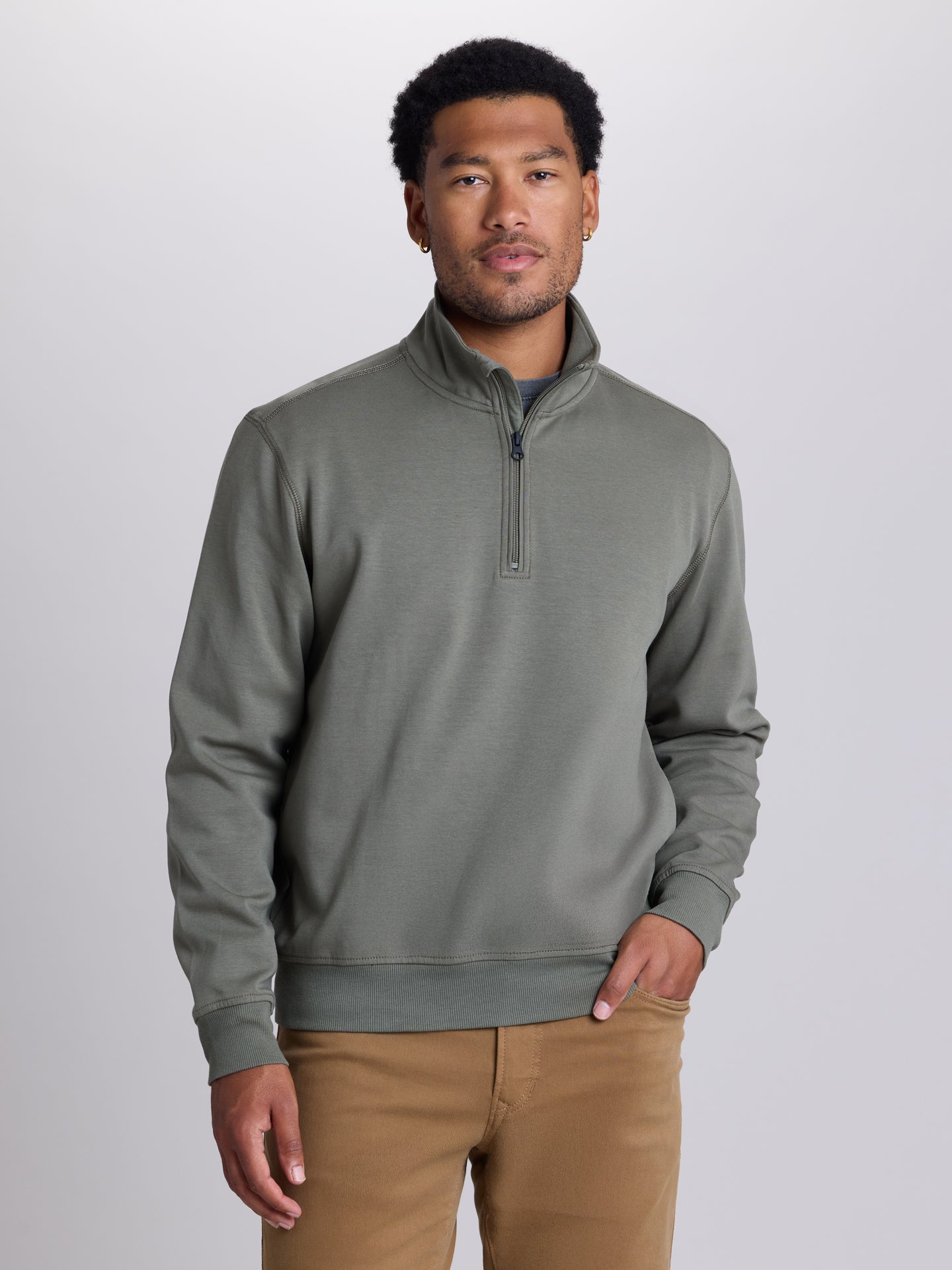 A man wearing a sage green quarter-zip pullover sweater and tan pants, standing against a light gray studio background. | Cobra
