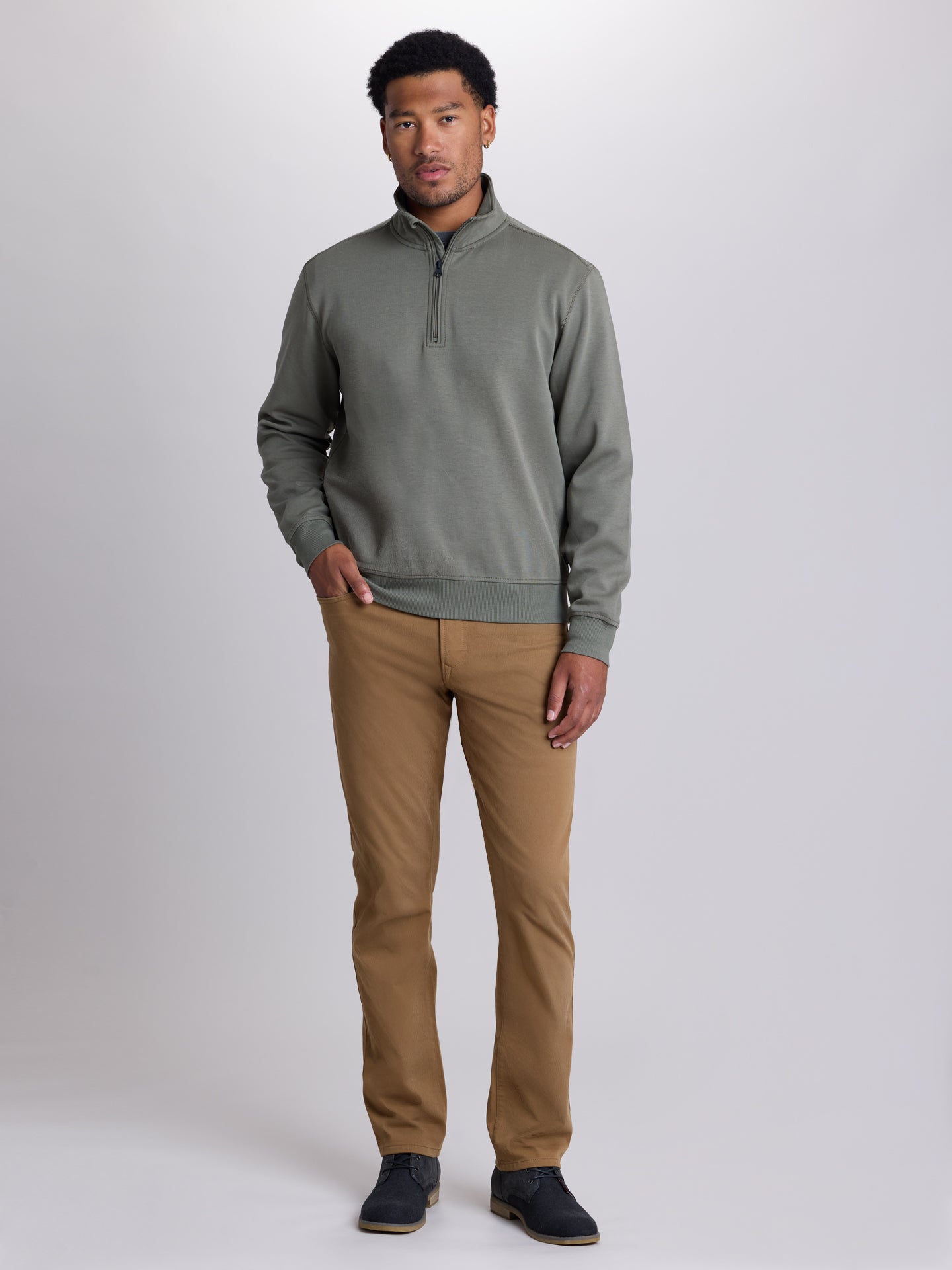 A man modeling a muted sage green quarter-zip pullover sweater paired with tan pants and dark blue shoes, standing against a light gray studio background. | Dusty Olive