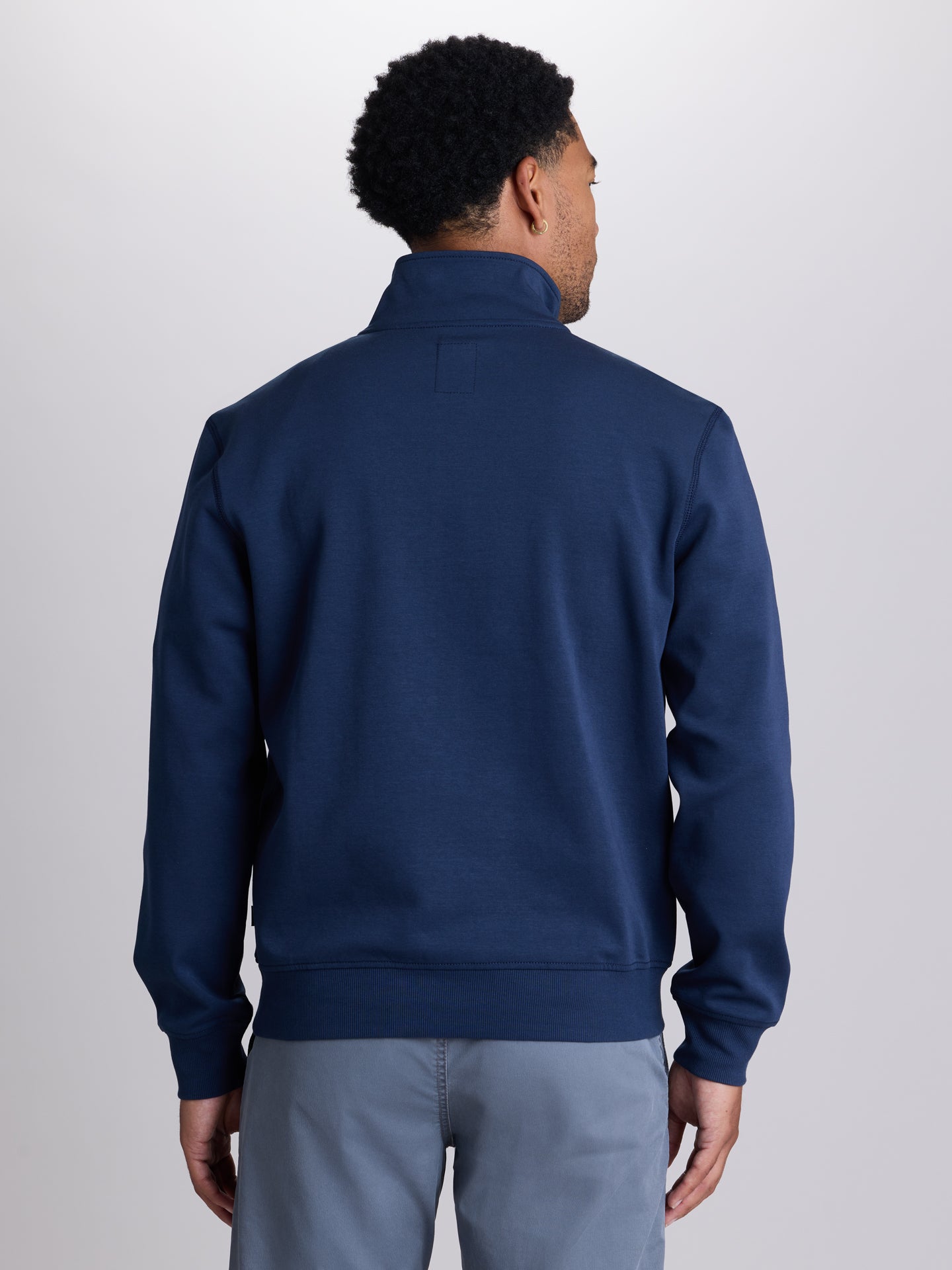 A man viewed from the back wearing a navy blue quarter-zip sweatshirt and light gray pants, set against a plain light gray studio background. | Starry