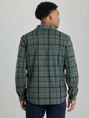 A man viewed from the back wearing a green and black plaid button-down shirt and navy pants, set against a plain light gray studio background. | Fog