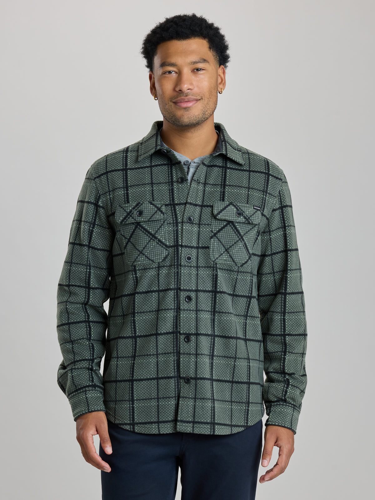 A man wearing a dark green and black plaid button-up shirt jacket over a light blue undershirt, paired with dark navy pants, standing against a solid light gray background. | Fog
