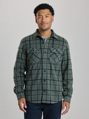 A man wearing a dark green and black plaid button-up shirt jacket over a light blue undershirt, paired with dark navy pants, standing against a solid light gray background. | Fog