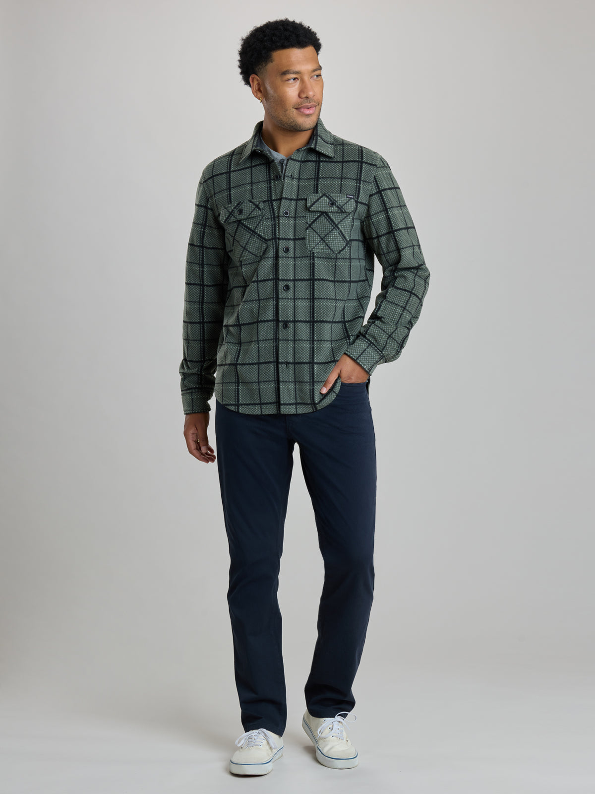 A man wearing a dark green and black plaid button-up shirt jacket over a light blue undershirt, paired with dark navy pants, standing against a solid light gray background. Full Shot of image. | Fog