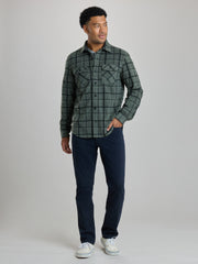 A man wearing a dark green and black plaid button-up shirt jacket over a light blue undershirt, paired with dark navy pants, standing against a solid light gray background. Full Shot of image. | Fog