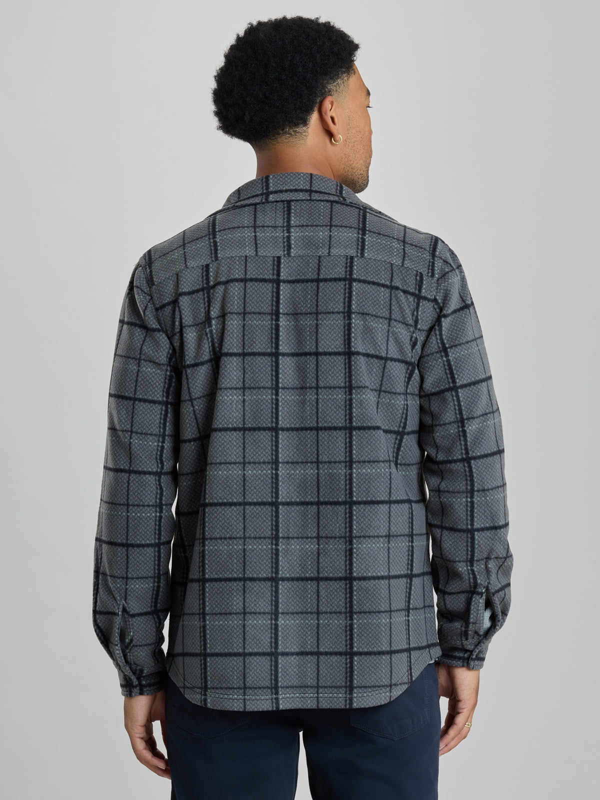 A man viewed from the back wearing a long-sleeve button-up shirt with a gray and black windowpane plaid pattern, paired with dark navy pants, against a solid light gray studio background.  | Forged
