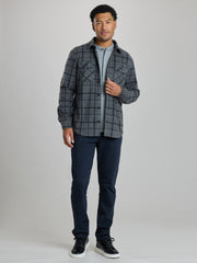 A man modeling a dark gray and black plaid button-up overshirt over a light gray t-shirt, paired with navy blue pants and black sneakers, against a solid light gray studio background. | Forged