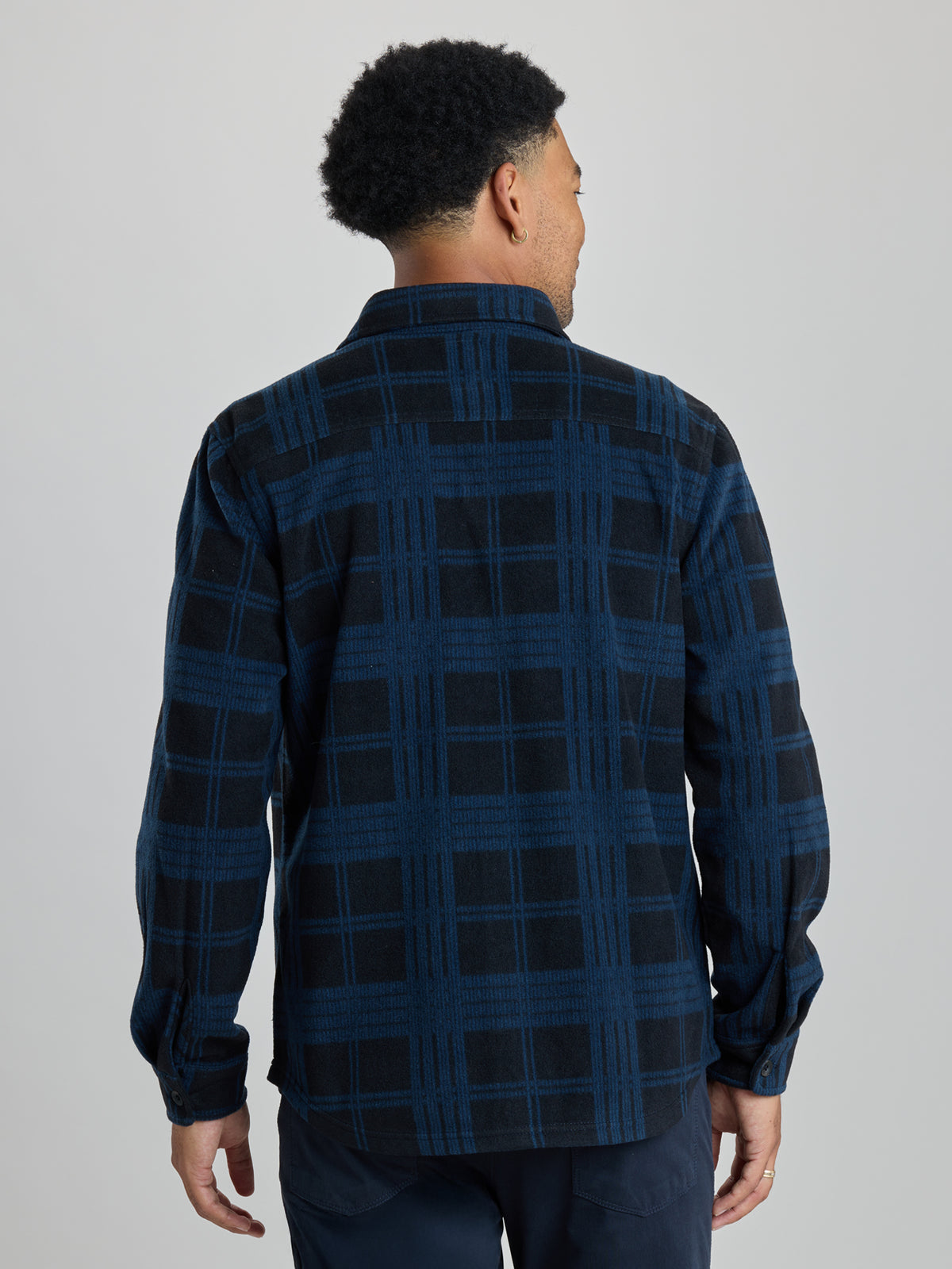 A man viewed from the back wearing a dark navy and black plaid long-sleeve shirt, paired with dark trousers, against a solid light gray studio background. | True Navy