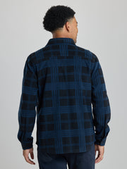 A man viewed from the back wearing a dark navy and black plaid long-sleeve shirt, paired with dark trousers, against a solid light gray studio background. | True Navy