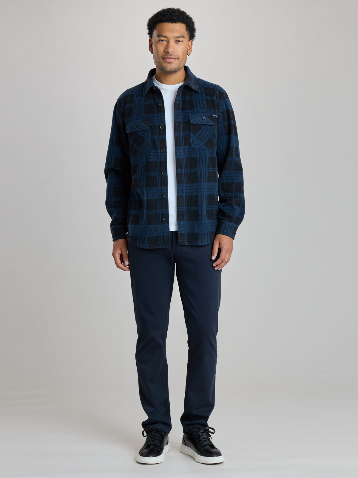 A man modeling a dark blue and black plaid button-up flannel shirt over a white t-shirt, paired with dark navy pants and black sneakers, against a light gray studio background. | True Navy