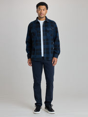 A man modeling a dark blue and black plaid button-up flannel shirt over a white t-shirt, paired with dark navy pants and black sneakers, against a light gray studio background. | True Navy